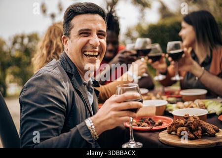 Ritratto sincero di un uomo sorridente che gusta del vino durante una cena all'aperto con gli amici. Atmosfera calda, emozioni reali, stile di vita autentico e allegro Foto Stock