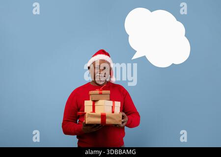 Uomo più anziano che indossa un cappello di Babbo Natale con scatole regalo davanti alla parete blu con la bolla vocale Foto Stock