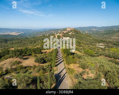 Vista aerea di una strada tortuosa costeggiata da alti e sottili alberi che conduce a un villaggio in cima a una collina sotto un cielo limpido, Montegiovi, Toscana, Italia. Foto Stock