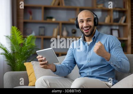 Il giovane uomo calvo dalla pelle scura con le cuffie si siede su un divano, regge un tablet e celebra la vittoria del gioco con un allegro pugno e un grande sorriso a casa Foto Stock