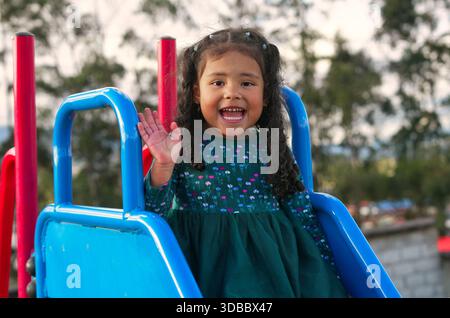 Sorridendo, in età prescolare, sventolando con gioia la mano mentre scivoli su uno scivolo blu vibrante in un parco giochi, per divertirsi all'aperto con una luminosa, S. Foto Stock