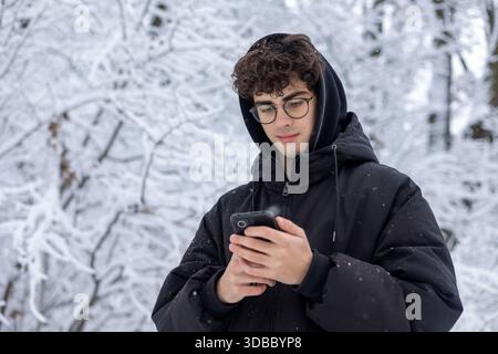 Ritratto di un teenager concentrato che usa lo smartphone all'aperto nello Snowy Winter Park. Giovane che controlla i social media o invia SMS, indossa occhiali e Black PUF Foto Stock