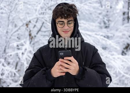 Ritratto di un teenager concentrato che usa lo smartphone all'aperto nello Snowy Winter Park. Giovane che controlla i social media o invia SMS, indossa occhiali e Black PUF Foto Stock