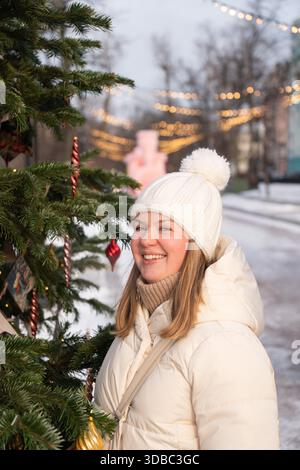 Una giovane donna sorridente con una giacca bianca invernale e un cappello a maglia si erge all'aperto vicino a un albero di Natale con calde luci cittadine sullo sfondo Foto Stock