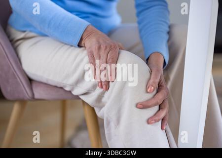 Donna anziana seduta su una sedia che tiene il suo doloroso ginocchio con entrambe le mani, sperimentando disagio e problemi di salute spesso associati a invecchiamento, artrite o lesioni Foto Stock