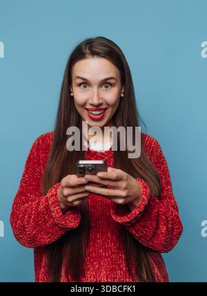 Una donna con i capelli lunghi regge uno smartphone e sorride con entusiasmo. Sembra essere al chiuso, con uno sfondo blu pieno e un'illuminazione brillante. Foto Stock