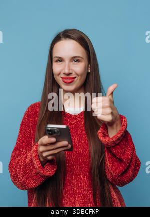 Donna in piedi con i capelli lunghi e il rossetto rosso mentre tiene in mano uno smartphone. Sorride e mostra un pollice in su. Lo sfondo è blu. Foto Stock