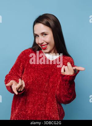 Una donna con lunghi capelli scuri si trova di fronte a uno sfondo blu. Indossa un maglione rosso e ha un sorriso brillante mentre fa giocosi gesti della mano Foto Stock