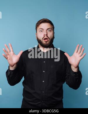 Un uomo sta con le mani in alto, mostrando la sua sorpresa. Ha un'espressione scioccata. Lo sfondo è blu. Quell'uomo indossa una camicia nera. Foto Stock