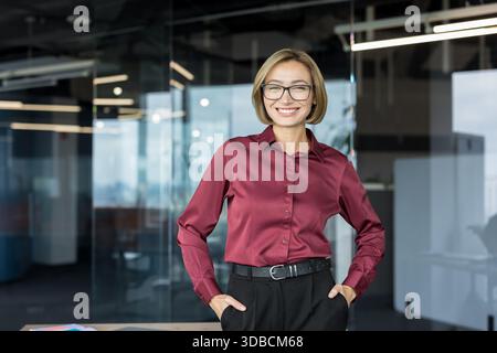 Giovane donna professionista che indossa occhiali da vista e una camicia marrone, sorridente con le mani in tasca, in piedi in un ambiente aziendale contemporaneo Foto Stock