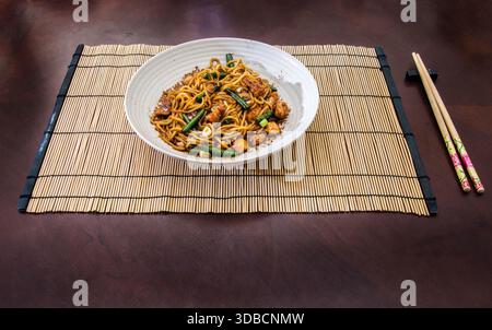 Una ciotola di spaghetti fritti al pollo serviti su un tappeto di bambù con bacchette, con verdure colorate e una presentazione di ispirazione asiatica. Foto Stock