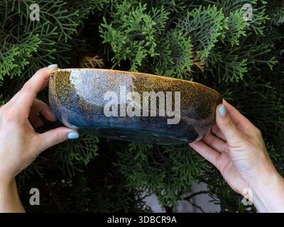 Recipiente in ceramica realizzato a mano con glassa blu e marrone ruvida tenuta in mani femminili su sfondo verde fogliame. Immagini di prodotti con luce naturale in alto Foto Stock