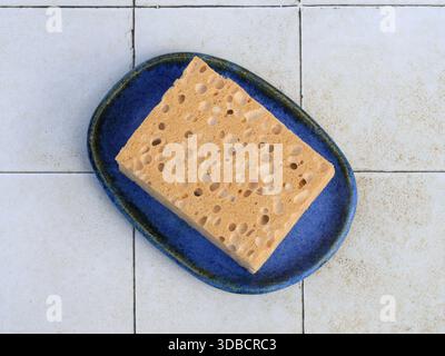 Vista dall'alto di una spugna porosa naturale da bagno poggiata su una teglia di sapone in ceramica smaltata blu su una superficie piastrellata. Il bagno era pulito e minimalista, con particolare attenzione al design su Foto Stock