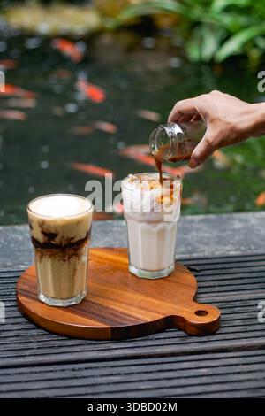 Caffè ghiacciato e latte ghiacciato serviti su tavola di legno, sciroppo di caffè versato, atmosfera da caffetteria all'aperto Foto Stock