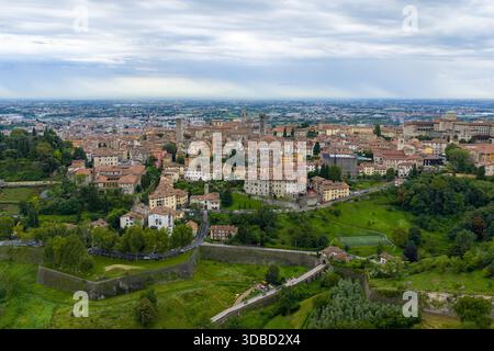 Vista sopraelevata sui quartieri periferici di Bergamo e sui terreni agricoli sotto il cielo estivo coperto, con densi tetti di terracotta e verdi colline circostanti. Foto Stock