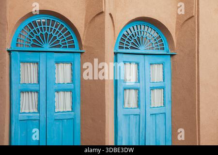 Due porte identiche in legno blu brillante con finestre decorative ad arco sono poste in una parete con texture marrone Foto Stock