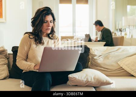 La moglie di mezza età si siede sul divano usando un laptop a casa, mentre sembra preoccupata per le finanze familiari e le banche online mentre suo marito lavora nel backgrou Foto Stock