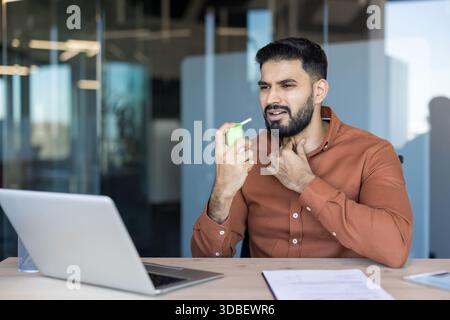 Giovane in un ufficio moderno si sente male con mal di gola, usando spray per la gola medicata mentre lavora su un computer portatile, mostrando malattia, autotrattamento e disagio sul posto di lavoro Foto Stock