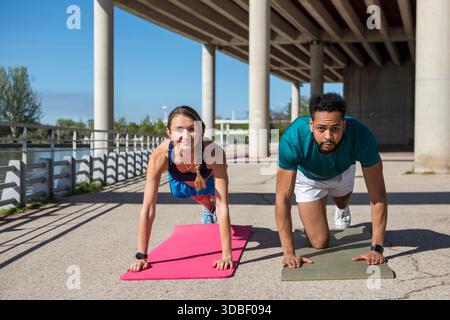 Due amici si allenano all'aperto sotto un ponte, facendo arrampicatori su tappetini da yoga. Il sole splende brillantemente, creando un'atmosfera vivace Foto Stock