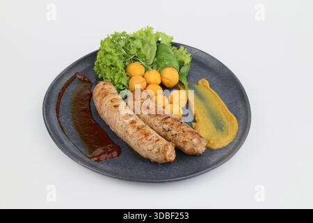 Salsicce grigliate con lattuga verde e crocchette di patate su un piatto scuro su sfondo bianco. Foto Stock