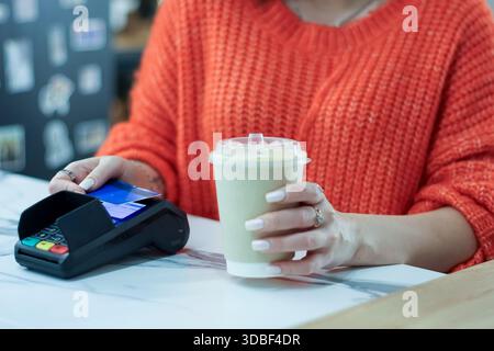 Primo piano della mano femminile inserendo la carta di credito nel terminale POS accanto alla bevanda cremosa in una tazza monouso su una superficie in marmo. Foto Stock