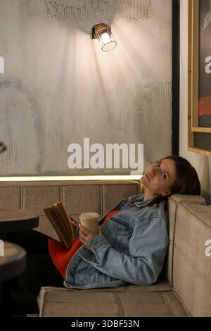 Giovane donna rilassata sul divano del caffè in giacca di denim che guarda attentamente verso l'alto mentre tiene un libro aperto e una tazza di caffè da asporto Foto Stock