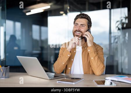 Giovane che ha una piacevole conversazione telefonica mentre è seduto alla sua scrivania, lavora su un notebook in un ambiente d'ufficio moderno, sorride e ha avuto successo nelle comunicazioni aziendali Foto Stock