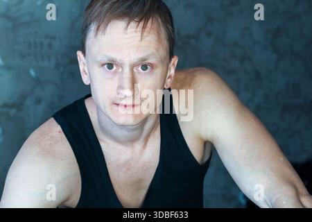 Uomo atletico dagli occhi larghi che si piega in avanti con un'espressione sconvolgente, indossa una camicia nera senza maniche in interni Foto Stock