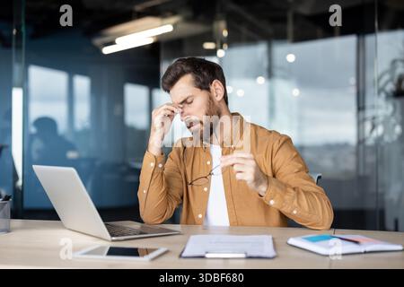 Un manager maschile stressato che tiene gli occhiali e sfrega le tempie, mentre soffre di un forte mal di testa e affaticamento degli occhi durante il lavoro sul suo laptop, in un ufficio moderno Foto Stock