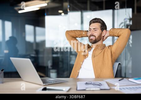 Giovane imprenditore felice che si prende una pausa dal lavoro, seduto sulla sedia con le mani dietro la testa, godendo del comfort e del successo in un ambiente di ufficio moderno Foto Stock