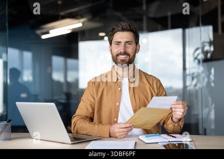 Un uomo d'affari sorridente a una scrivania moderna apre una busta marrone per rivelare un documento importante, celebrando buone notizie - successo professionale, approvazione o una corrispondenza positiva Foto Stock