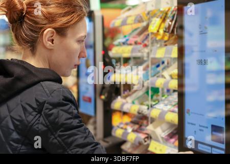 Profilo della donna dai capelli rossi che seleziona gli articoli sul distributore automatico touchscreen in negozio Foto Stock