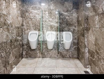 Bagno pubblico minimalista per uomini con tre orinatoi bianchi separati da pareti in vetro smerigliato contro pareti in marmo e pavimento piastrellato, moderno e pulito Foto Stock