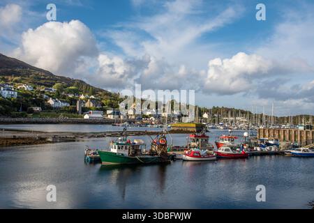 Una tranquilla scena portuale a Tarbert sul Loch Fyne, che mostra colorate barche da pesca, una piccola cittadina di mare e un cielo luminoso e parzialmente nuvoloso sull'acqua calma Foto Stock