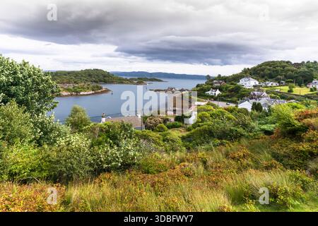 Una tranquilla scena costiera di Tarbert sul Loch Fyne, con case bianche, barche, giardini lussureggianti e una costa rocciosa. Colline ondulate, un cielo nuvoloso e.. Foto Stock