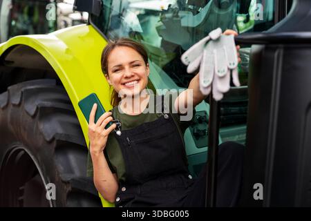 Allegra lavoratrice agricola con smartphone in piedi vicino al trattore agricolo. Foto Stock