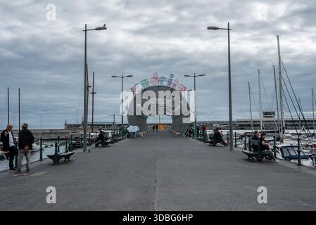 Un ampio molo a Funchal, Madeira conduce a un arco festoso circolare con stelle e nuvole. Le persone si siedono sulle panchine verdi mentre le barche a vela fiancheggiano il porticciolo sotto Foto Stock