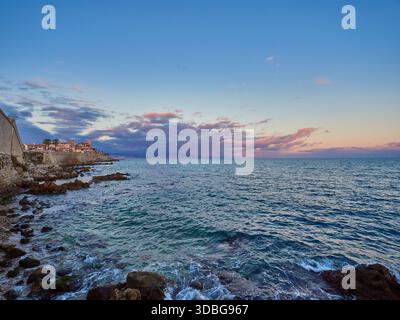 Tramonto costiero sul mare calmo con le storiche mura di pietra e la costa rocciosa con la città costiera di Antibes, Francia Foto Stock