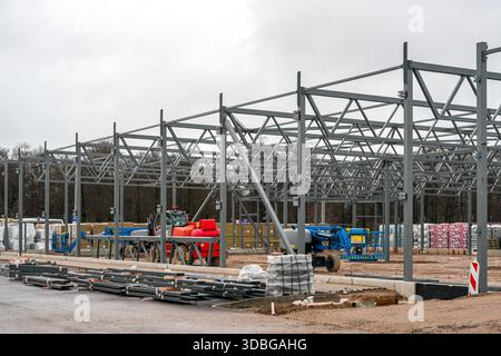 Grande cantiere industriale che mostra l'assemblaggio di un magazzino con telaio in acciaio con gru, sollevatori aerei e materiali da costruzione preparati per l'installazione Foto Stock