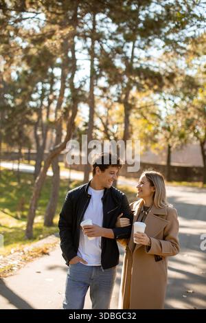 Due persone passeggiano insieme in un parco durante l'autunno. Si stanno gustando un caffè e sorridendo l'un l'altro. Gli alberi li circondano, mostrando giallo e arancione Foto Stock