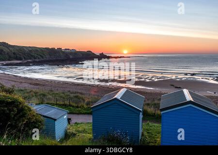 Tranquillo scenario costiero a Coldingham Bay con capanne blu sulla spiaggia, onde dolci e un vivace tramonto. Foto Stock