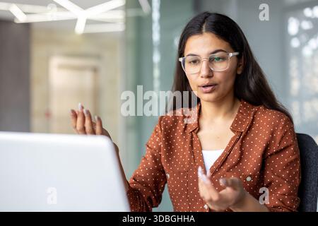 Giovane donna d'affari con occhiali trasparenti che partecipa attivamente a una videochiamata online. Comunicare e discutere le strategie aziendali lavorando in remoto da un ambiente di ufficio moderno Foto Stock