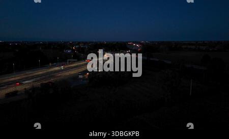 Vista aerea notturna dell'autostrada A21 con corsie illuminate, auto e camion in movimento attraverso un complesso interscambio e una piazza a pedaggio, mostrando un traffico intenso Foto Stock