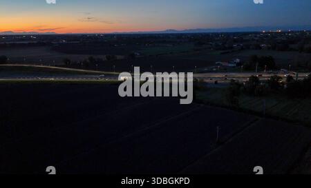 Il traffico dell'autostrada A21 che scorre di notte sull'autostrada Brescia Piacenza, auto e camion si spostano lungo una strada illuminata con uno skyline urbano e una foce Foto Stock