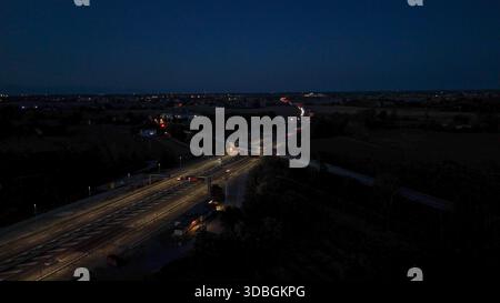 Vista aerea notturna dell'autostrada A21 con corsie illuminate, auto e camion in movimento attraverso un complesso interscambio e una piazza a pedaggio, mostrando un traffico intenso Foto Stock