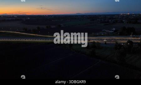 Il traffico dell'autostrada A21 che scorre di notte sull'autostrada Brescia Piacenza, auto e camion si spostano lungo una strada illuminata con uno skyline urbano e una foce Foto Stock