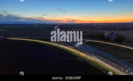 Vista aerea che monitora auto e camion che viaggiano sull'autostrada A21, un importante percorso di trasporto, con un cavalcavia illuminato dalla strada li Foto Stock