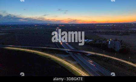 Autostrada A21 che mostra il flusso del traffico serale, veicoli che si muovono lungo l'autostrada illuminata e strade di cavalcavia che collegano città lontane sotto un bellissimo stato Foto Stock