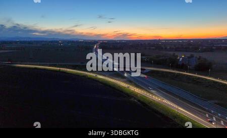 Vista aerea che monitora auto e camion che viaggiano sull'autostrada A21, un importante percorso di trasporto, con un cavalcavia illuminato dalla strada li Foto Stock