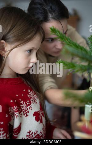 Ragazza in maglione rosso natalizio accanto alla madre che decora l'albero, accogliente vacanza in famiglia a casa Foto Stock